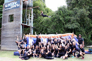 A large group of people smiling for the camera in front of an orange and blue airbag while one girl jumps from a seven meter high wooden platform onto the airbag.