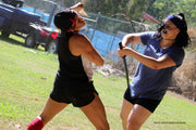 Two girls participating in a stunt sword fight. One girl is knocking the other in the face with the back of her sword.