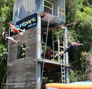Four people are participating in different activities on a wooden tower. Two of the four, a boy and a girl are abseiling down the tower. One boy is jumping face first off the tower to a large airbag from five meters high. One girl is patiently waiting for her turn to jump next.  