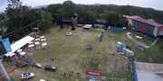 Aerial view of the Stunt Park facilities. The facilites include a large green lawn, wooden tower, and covered training area. In this photo an event is happening at stunt park and there are round tables lined up for food and drink. 