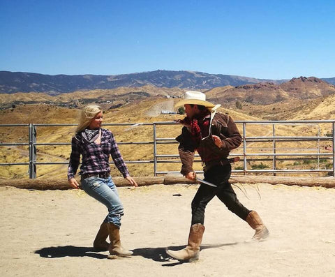Two people dressed in western apparel in the Hollywood mountains participating in a stunt fight. the boy has a plastic knife and the girl is dodging his swing. 