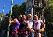 Four happy women in rock climbing harness stand together smiling as they prepare to abseil on a wooden tower. 