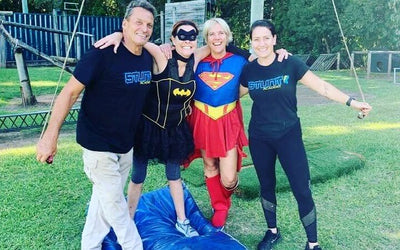 Three women and one man standing together and smiling for a photo. Two of the women are wearing super hero costumes. One is batman and one is superman